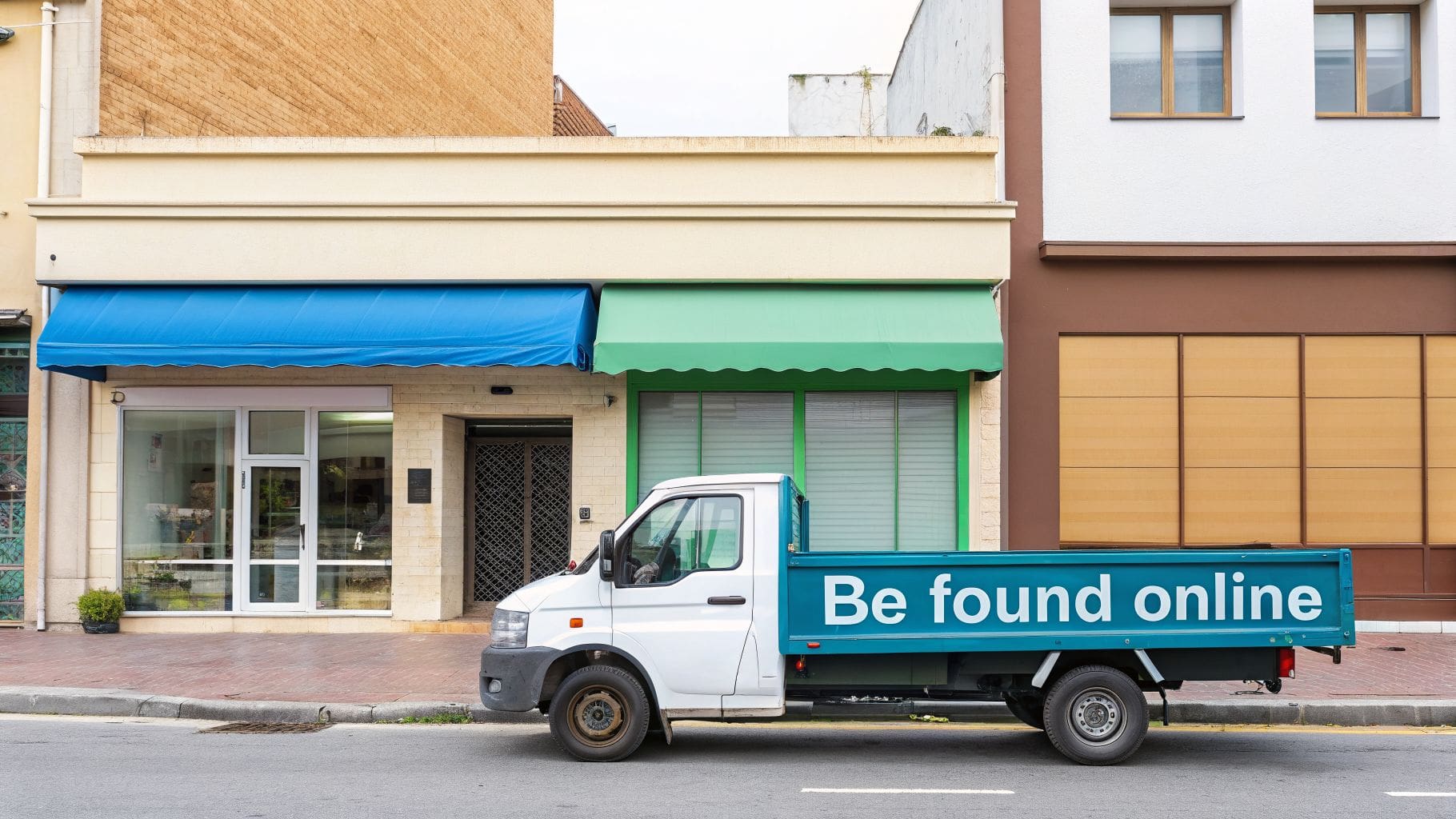 A white truck with 'Be found online' on its side is parked on a street in front of small businesses.